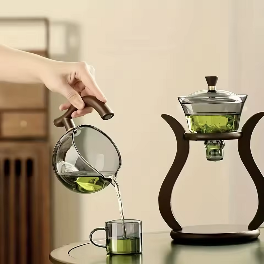 Hand pouring tea from lower glass pot into small cup beside brewing stand