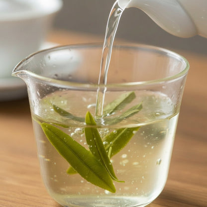 Tea leaves being poured into a glass cup with a teapot in the background.