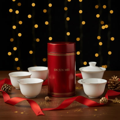 Red tea canister labeled 'JIN JUN MEI' surrounded by white teacups on a wooden surface with decorative lights in the background.